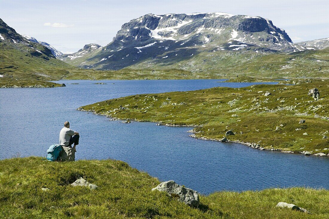 Man with backpacker is sitting on a stone a looking out on water and mountains, Hardangervidda, Norway