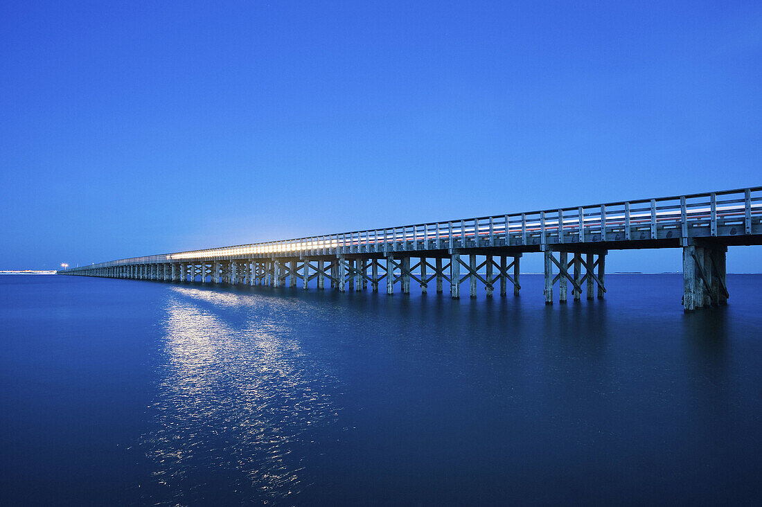 The lights of a vehicle reflect off the water as it travels over the half mile long Powder Point wooden bridge spanning a cove at dusk, Massachusetts, USA