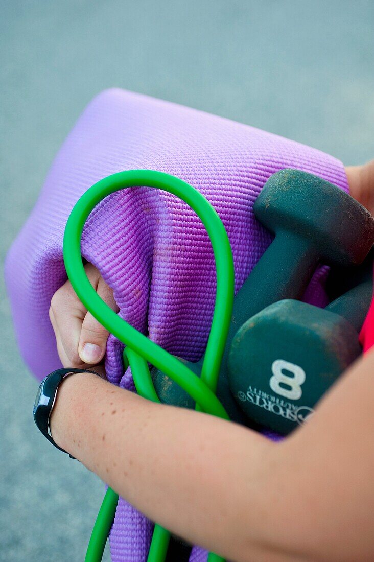 Two dumbbells, a yoga matt and a workout rubberband in the arms of a young woman.