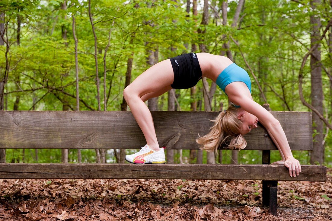 A young blond woman doing a yoga back bend posture on a wooden bench in a park.