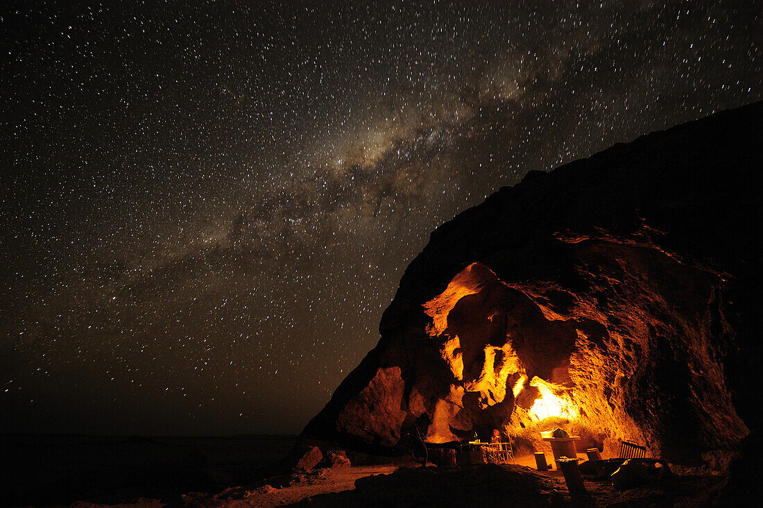 Personen sitzen bei Grillfeuer unter beleuchtetem Felsüberhang mit Sternenhimmel mit Milchstraße, Namib Naukluft National Park, Namibwüste, Namib, Namibia