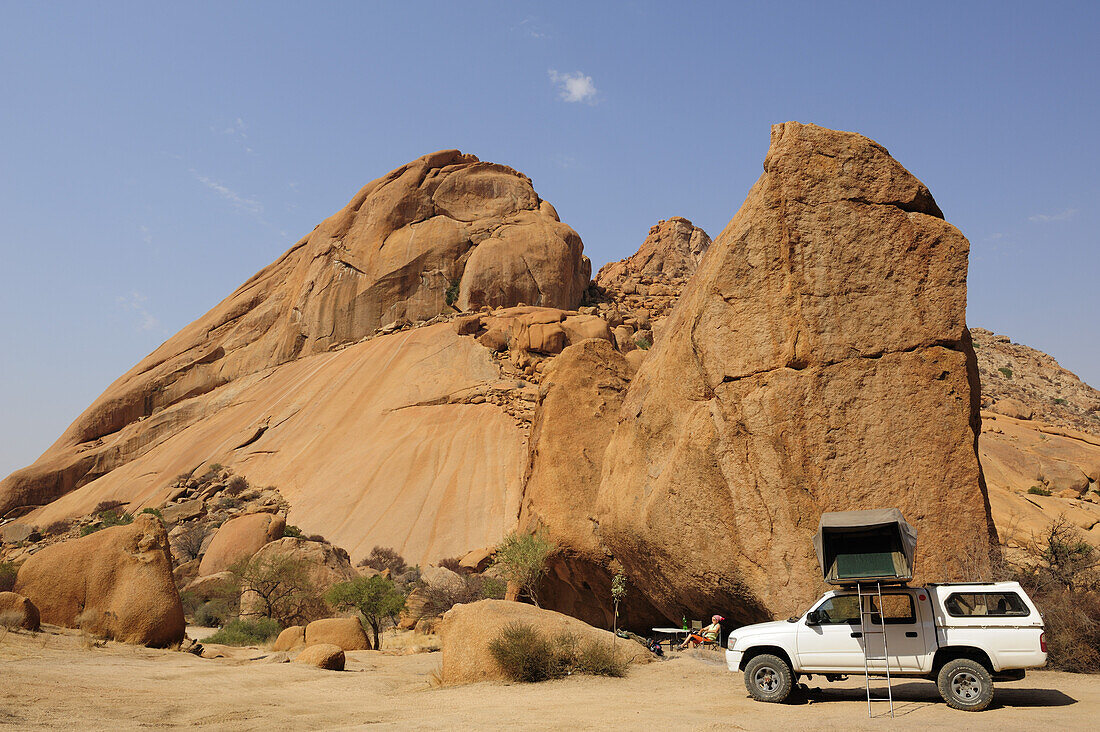Frau sitzt vor rotem Felsmassiv, Auto mit Dachzelt im Vordergrund, Große Spitzkoppe, Namibia