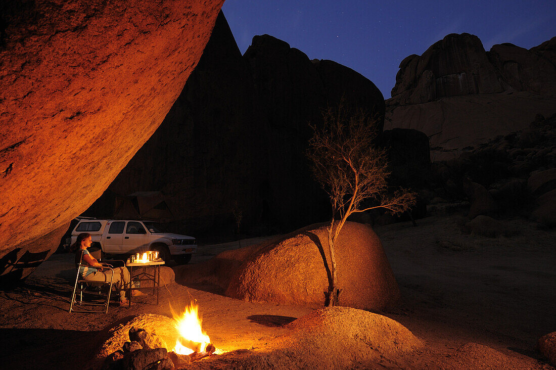 Woman sitting in front of barbecue fire under rock overhang, car in background, Great Spitzkoppe, Namibia
