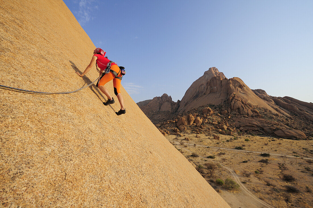 Frau klettert an roter Felswand, Große Spitzkoppe im Hintergrund, Große Spitzkoppe, Namibia