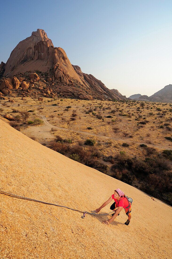 Frau klettert an roter Felsplatte, Große Spitzkoppe im Hintergrund, Große Spitzkoppe, Namibia