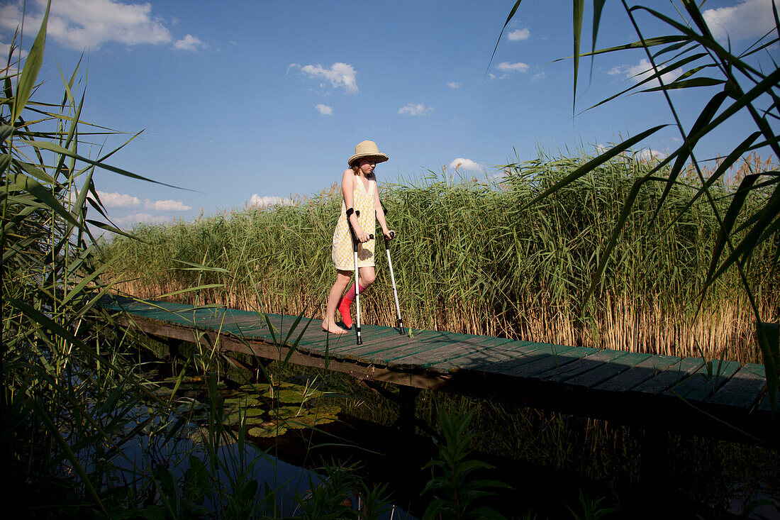 Mädchen mit Gipsbein geht auf Krücken über einen Steg, Teupitzer See, Brandenburg, Deutschland