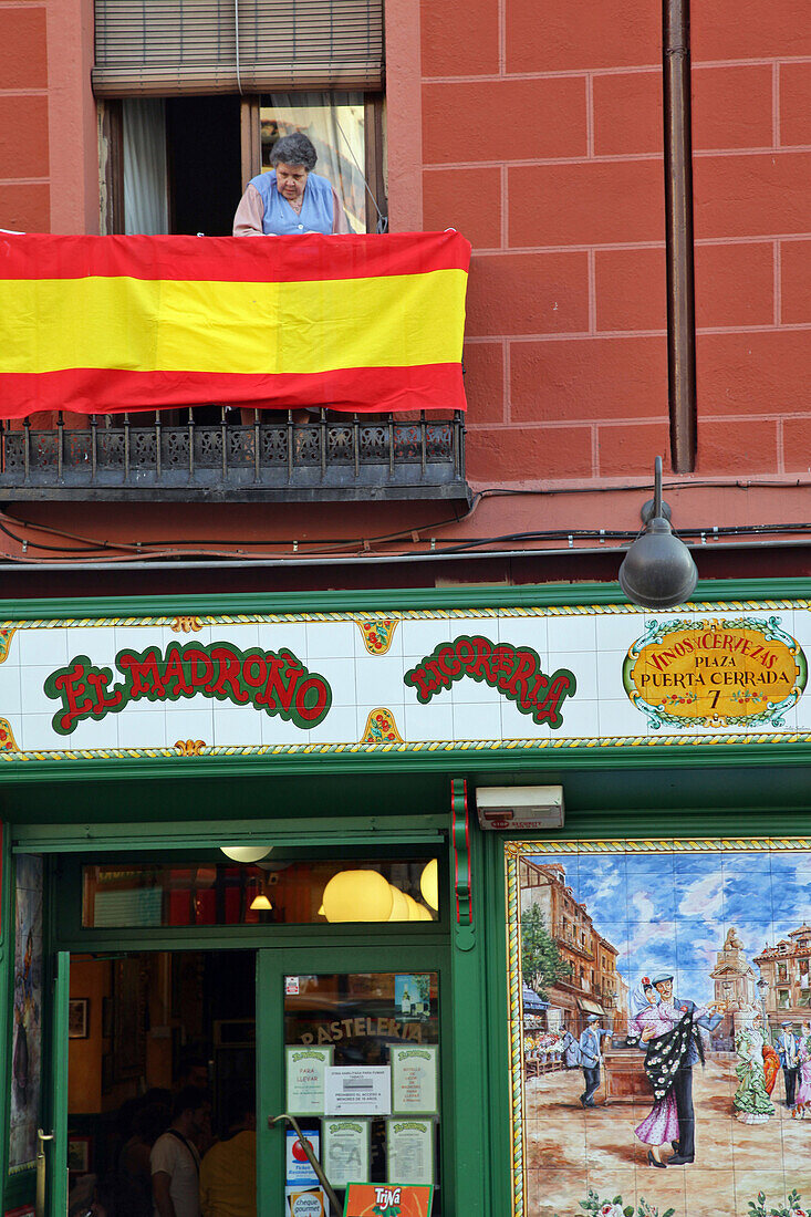 Old Woman With A Spanish Flag At Her Window Over The Restaurant El Madrono, Plaza Puerta Cerrada, Madrid, Spain