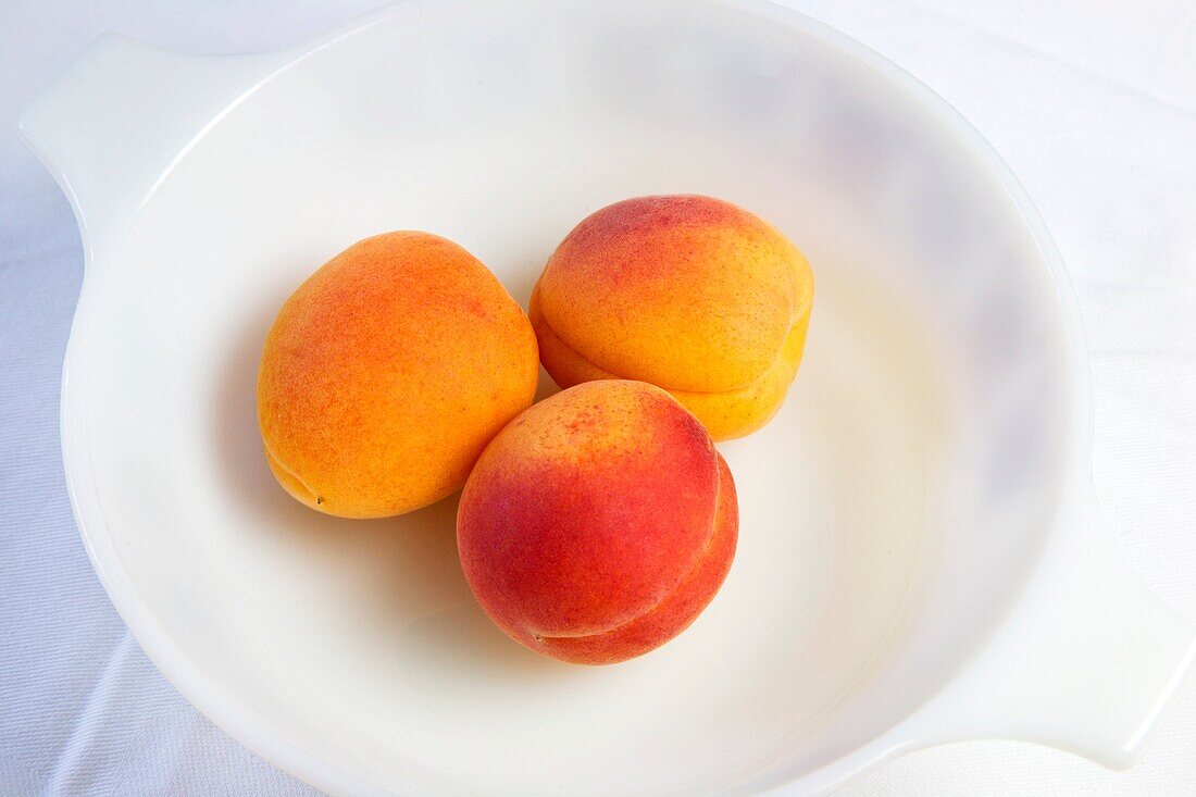 Three apricots in a white bowl on a white cloth