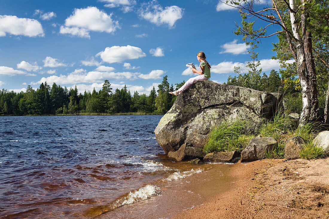 9 year old girl sitting on a big rock at St. Hindsjön lake in the sunlight, South Sweden, Scandinavia, Europe