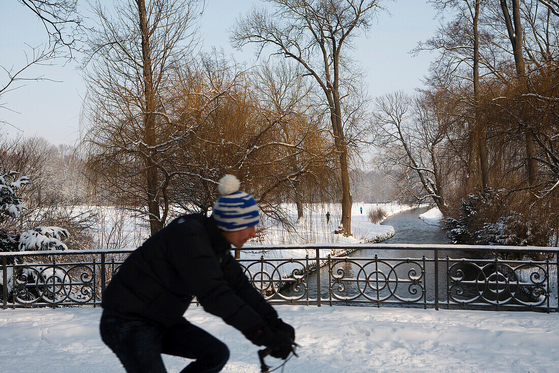 Cyclist passing bridge ober river Eisbach in winter, English Garden, Munich, Bavaria, Germany