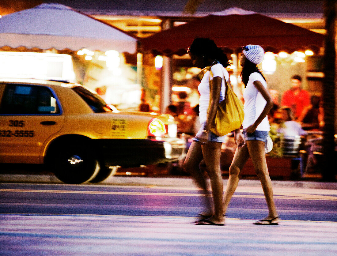 Two young women walking down the street at night in front of restaurants, Miami, Florida, USA
