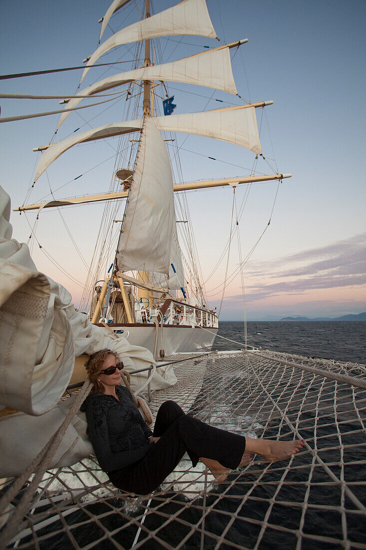 Junge Frau entspannt sich im Netz am Bugspriet vom Großsegler Kreuzfahrtschiff Star Flyer (Star Clippers Cruises) bei Sonnenuntergang, Pazifischer Ozean nahe Costa Rica, Mittelamerika, Amerika