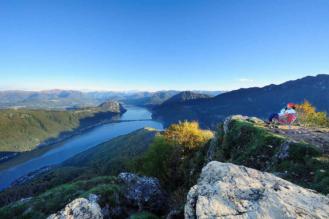 Woman sitting on red bench and looking towards lake Lugano, Ticino range in background, View from the  Monte San Giorgio, UNESCO World Heritage Site Monte San Giorgio, lake Lugano, Ticino range, Ticino, Switzerland, Europe