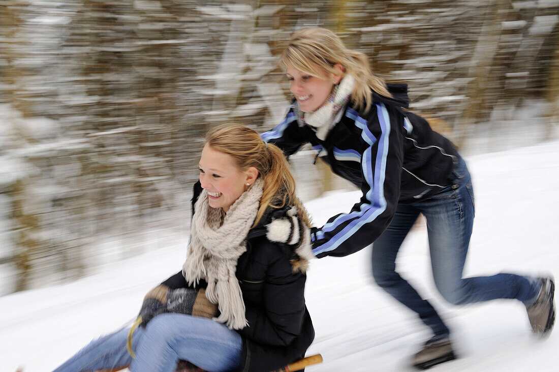 Young woman pushing another young woman on a sleigh, valley of Leitzachtal, Upper Bavaria, Bavaria, Germany, Europe