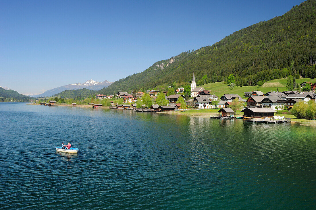 Two persons rowing a boat on lake Weissensee, village Gatschach in the background, lake Weissensee, Carinthia, Austria, Europe