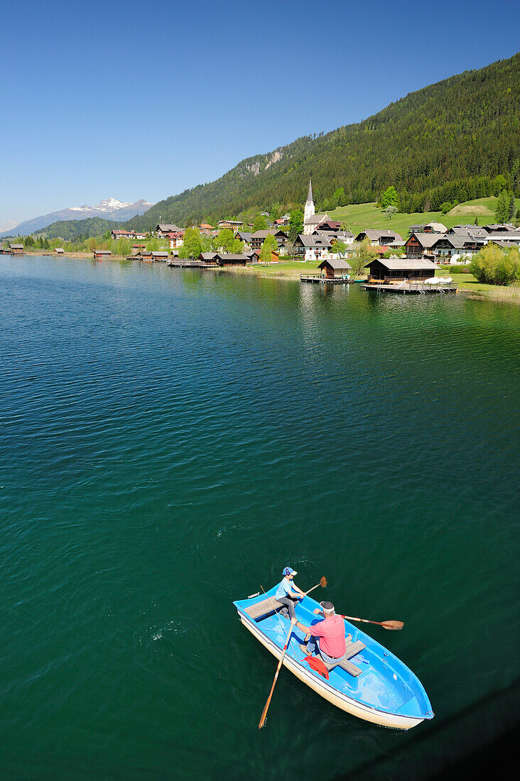 Two persons rowing a boat on lake Weissensee, village Gatschach in the background, lake Weissensee, Carinthia, Austria, Europe