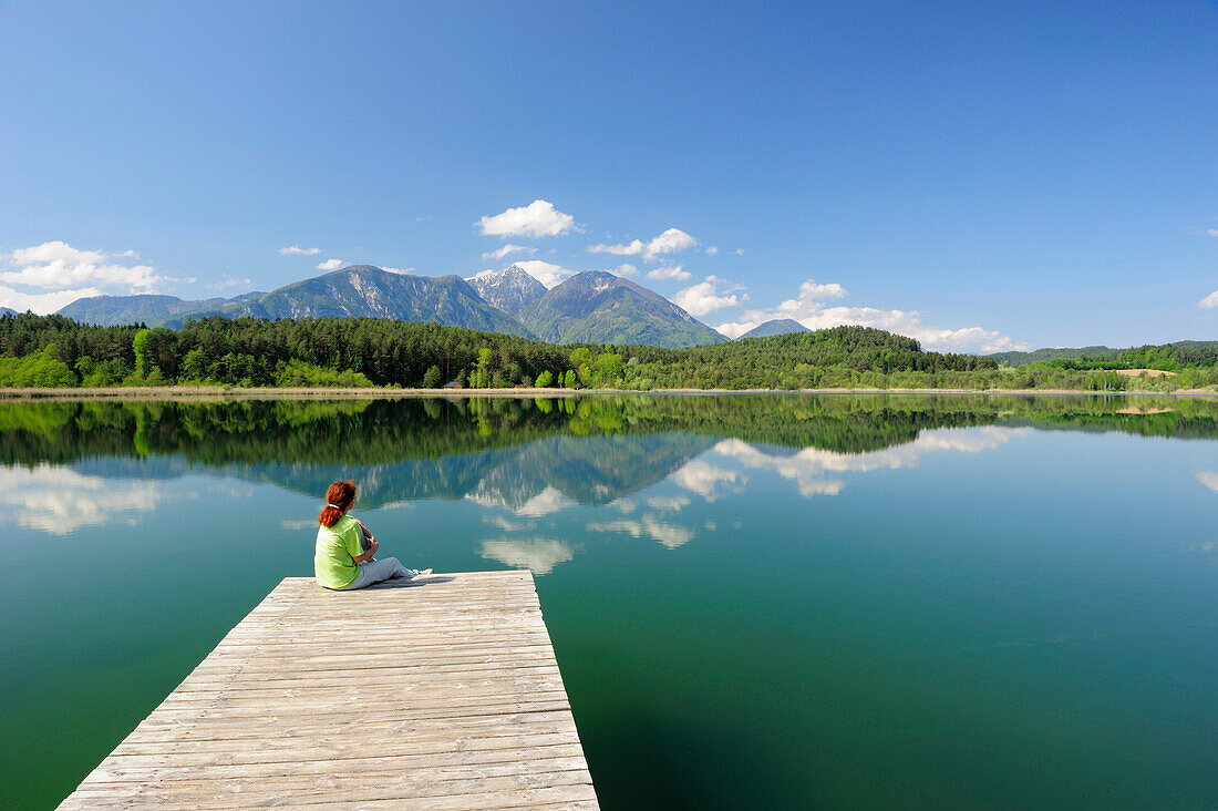 Frau sitzt auf Holzsteg am Turnersee und blickt auf Karawanken, Turnersee, Kärnten, Österreich, Europa