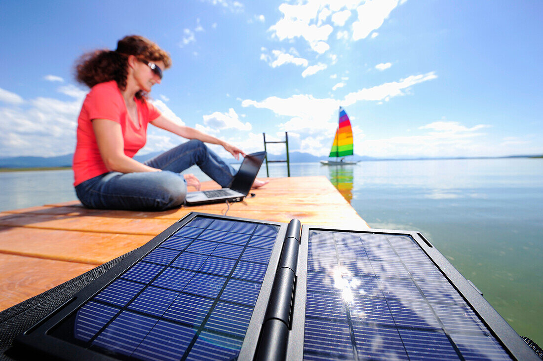 Woman using a laptop on a jetty at lake Chiemsee, solar panels in foreground, Chiemgau, Upper Bavaria, Bavaria, Germany