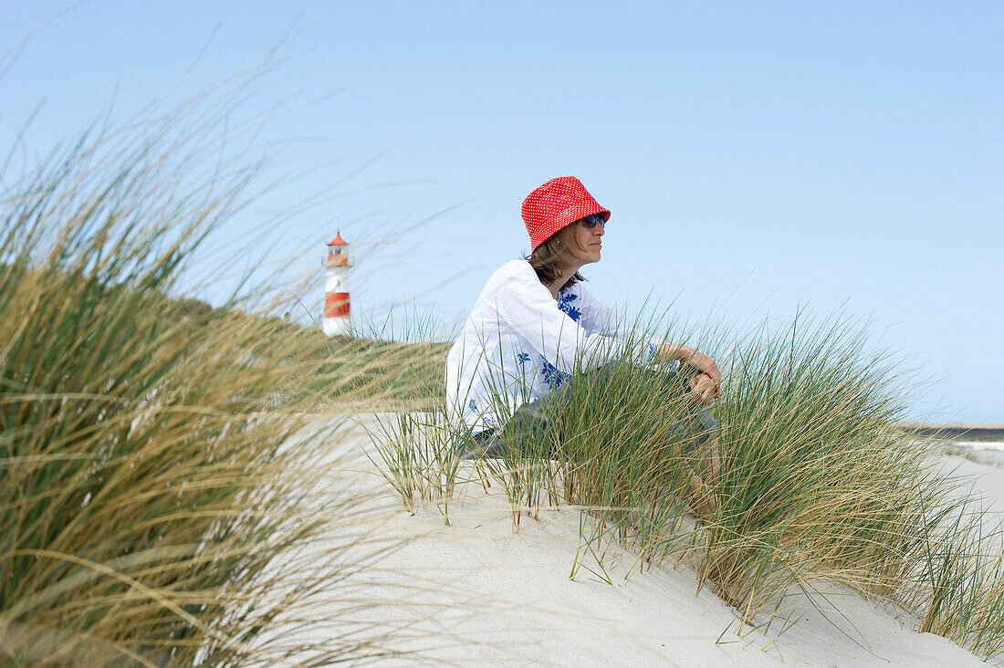 Woman wearing a red hut sitting at beach, List-Ost lighthouse in background, Ellenbogen, List, Sylt, Schleswig-Holstein, Germany