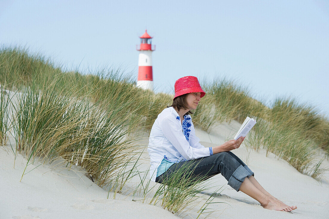 Frau mit Hut sitzt am Strand und liest, Leuchtturm List-Ost im Hintergrund, Ellenbogen, List, Sylt, Schleswig-Holstein, Deutschland