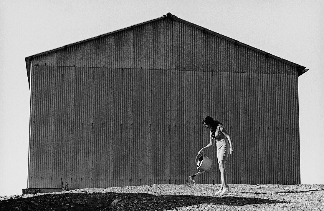 Woman watering ground with watering can behind warehouse, b&w