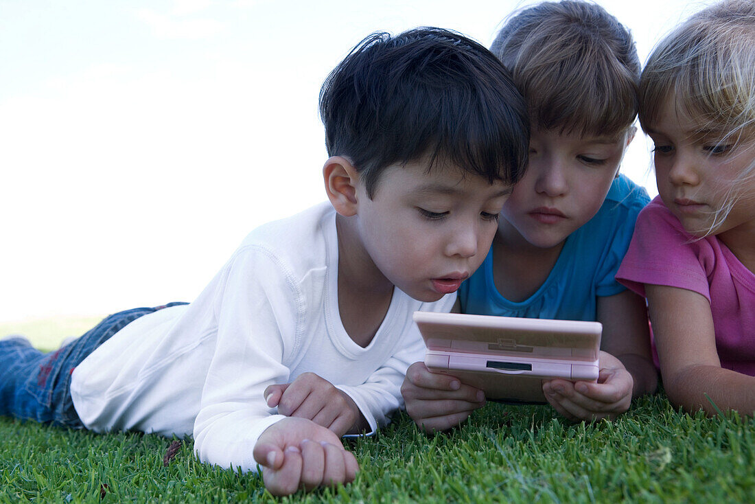 Three children lying on grass, playing with video game, close-up