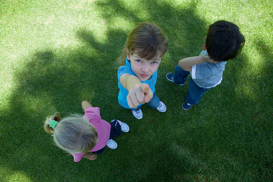 Three children standing on grass, in shade, one girl pointing at camera, view from directly above