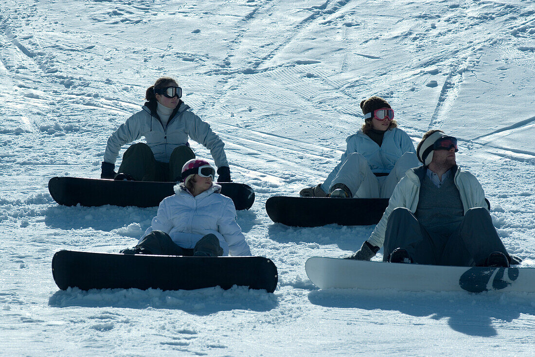 Four young snowboarders sitting on ski slope, looking away