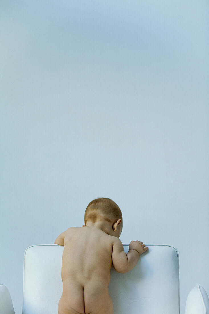 Nude baby standing on armchair, looking over the back, rear view