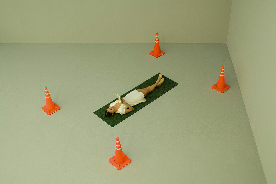 Woman lying on artificial turf and reading book, surrounded by traffic cones, high angle view