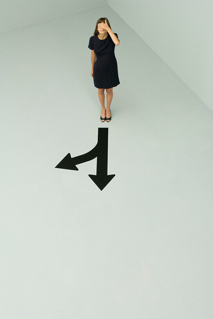 Woman standing before arrows pointing different directions, hand covering eyes, high angle view