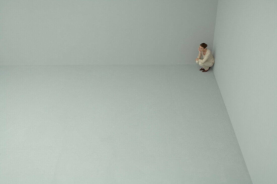 Woman crouching in corner of large empty room, mid-distance, high angle view