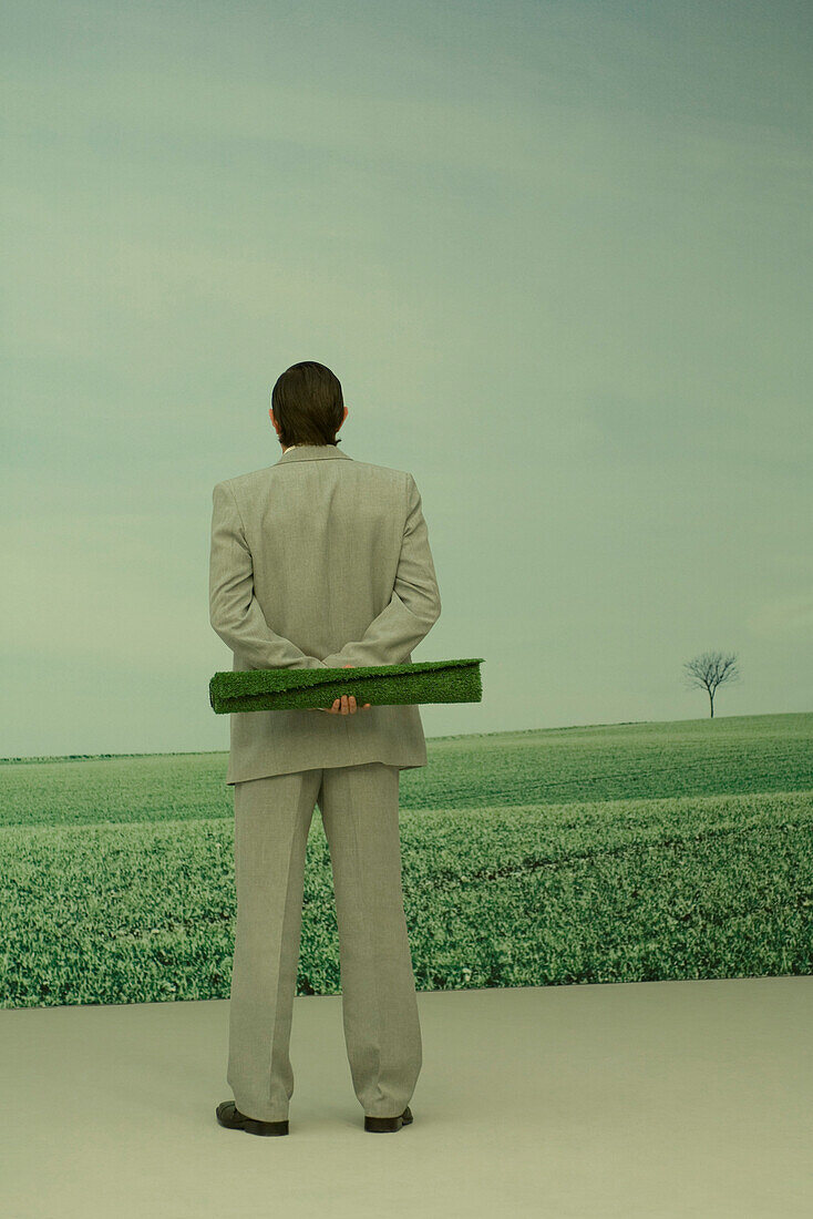 Man looking at rural scene, holding roll of artificial turf behind back, rear view