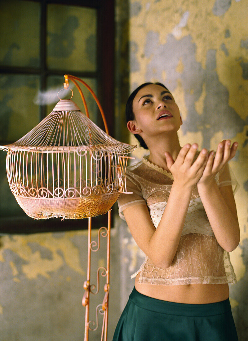 Woman standing beside empty birdcage with hands raised, feathers floating around her