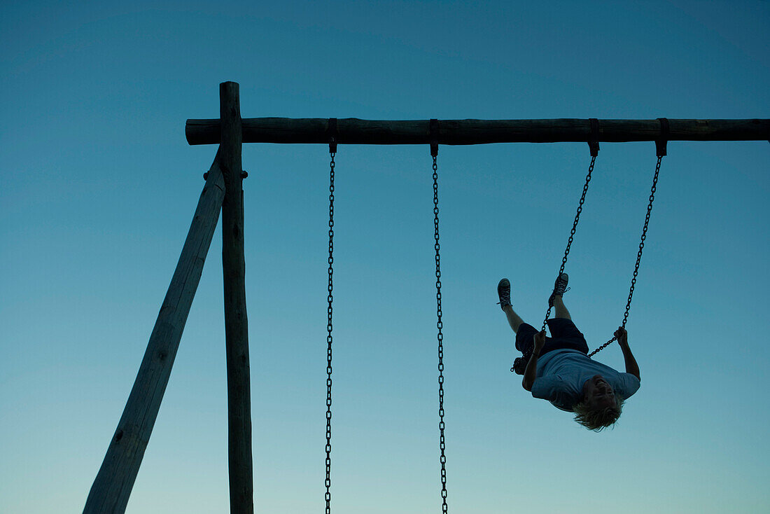 Young man swinging on swing set, backlit