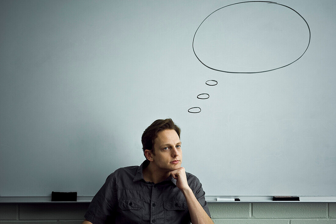 Teacher sitting in classroom contemplatively looking away, empty word bubble above his head