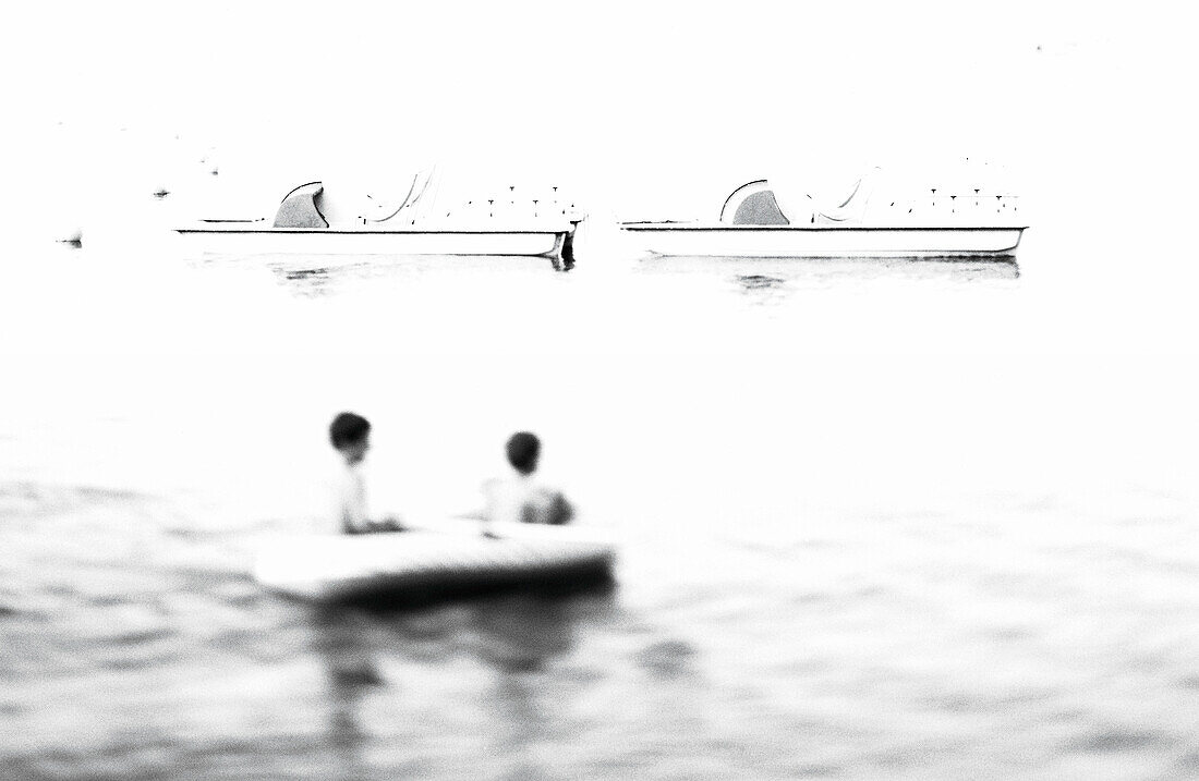 People on water in inflatable raft with pedal boats in background, b&w