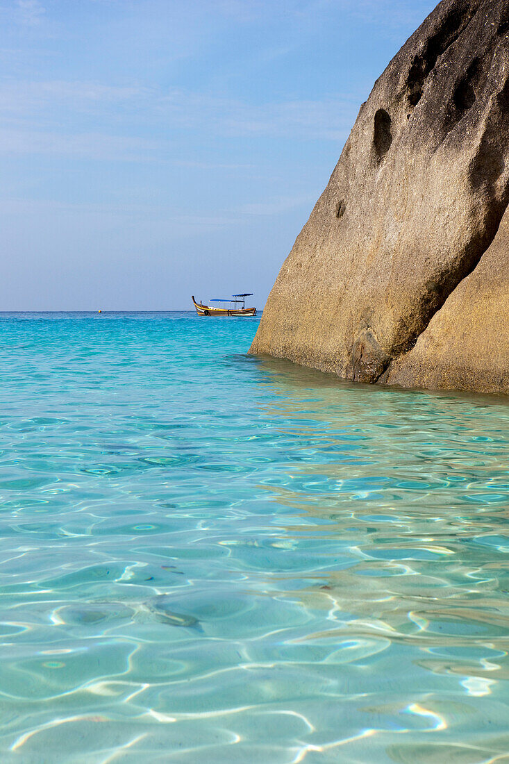 Glasklares Wasser umspühlt Felsen am Strand mit Longtailbooten im Hintergrund, Similan Inseln, Andamanensee, Thailand