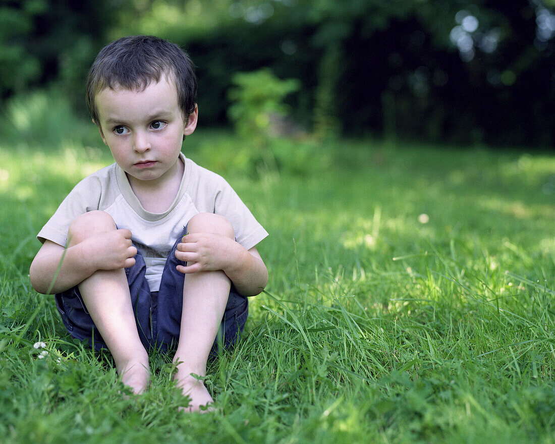 Little boy sitting in grass, looking away in thought