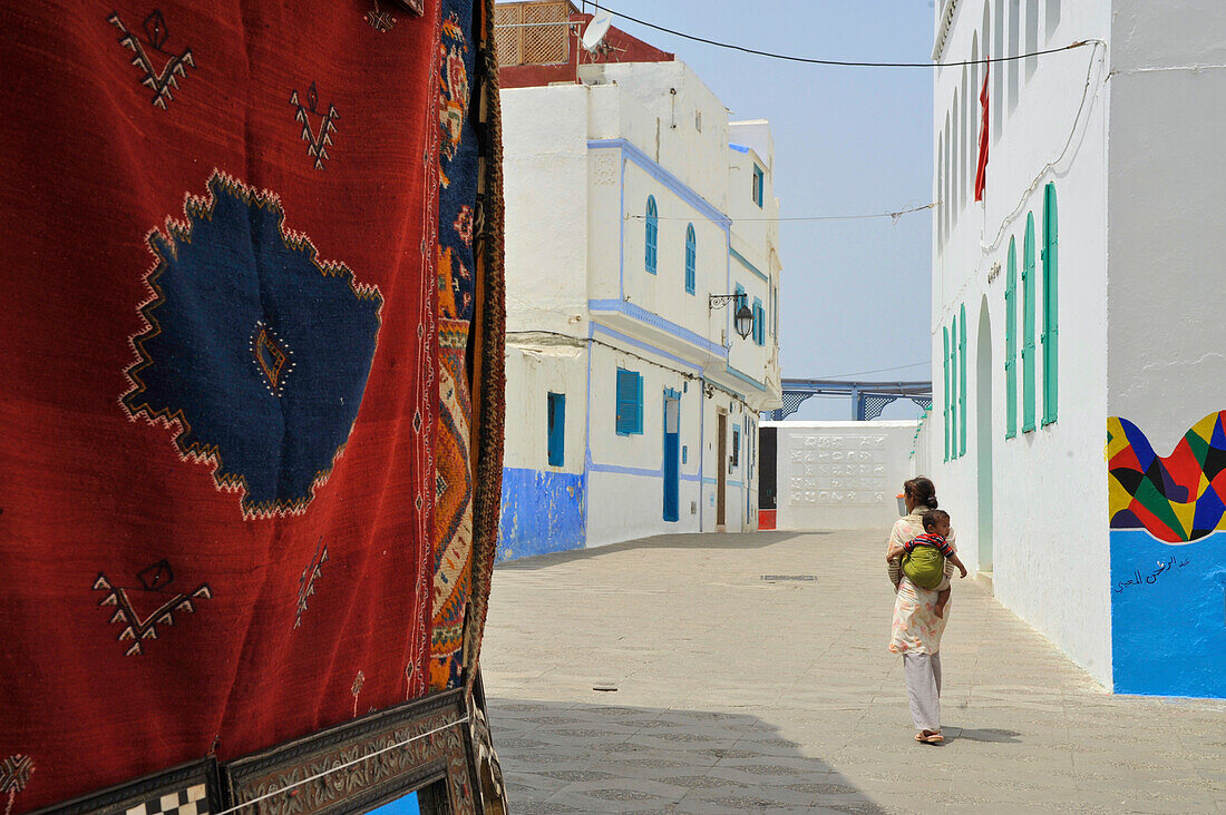 Teppich und bemalte Fassade und Frau mit Kind in der Altstadt in Asilah, Atlantik Küste, Marokko, Afrika