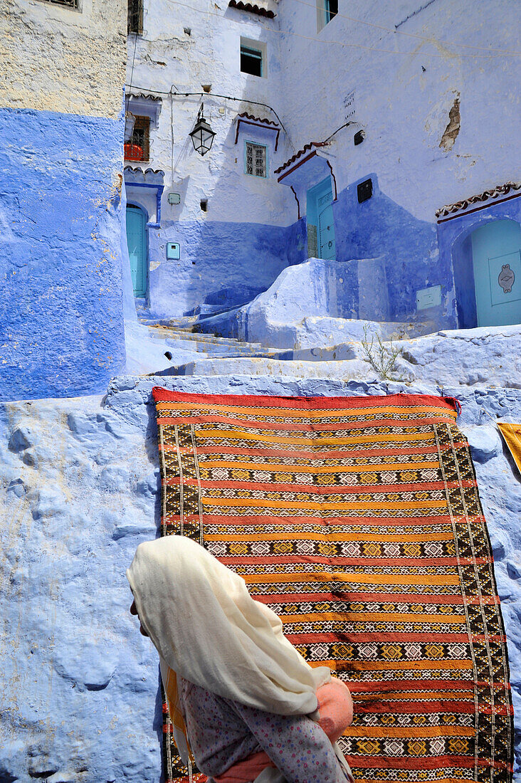 Alte Frau vor Teppich und Gasse mit blau bemalte Häusern in Chefchaouen, Riff Gebirge, Marokko, Afrika