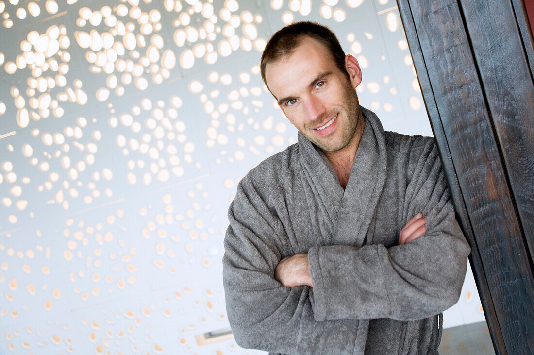 Man in grey bathrobe, leaning against wall, with crossed arms, smiling for the camera