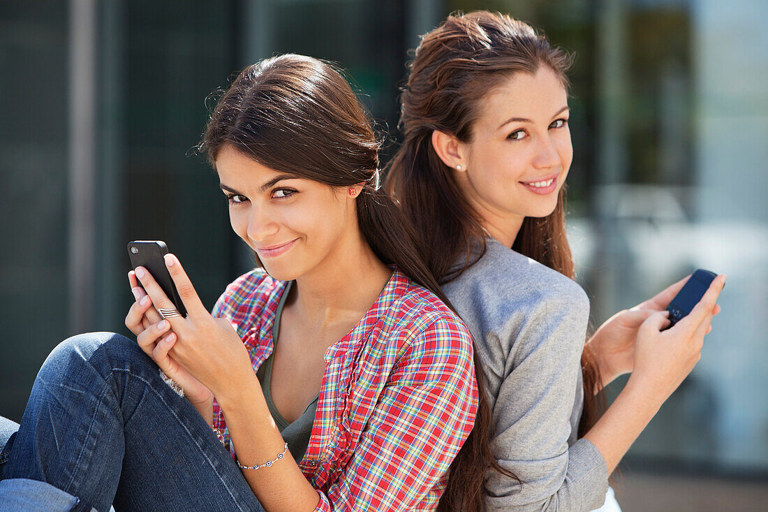 Portrait of two young women sitting back to back and using mobile phones