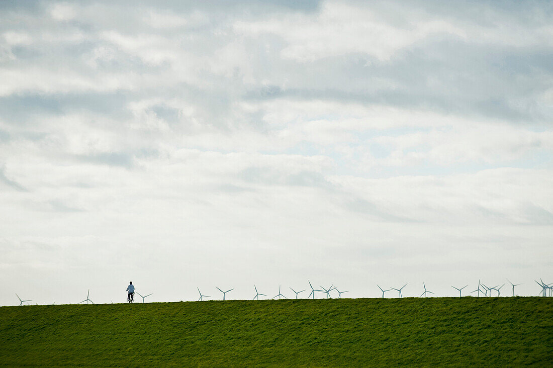 Female cyclist passing dike, wind turbines in background, Norderney, East Frisian Islands, Lower Saxony, Germany