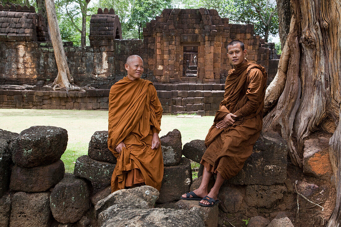 Zwei Mönche in braunem Gewand sitzen auf Mauer und neben einem alten Baum vor den Ruinen eines Khmer Tempel aus dem 13. Jahrhundert am Muang Singh Historical Park, nahe Kanchanaburi, Thailand, Asien