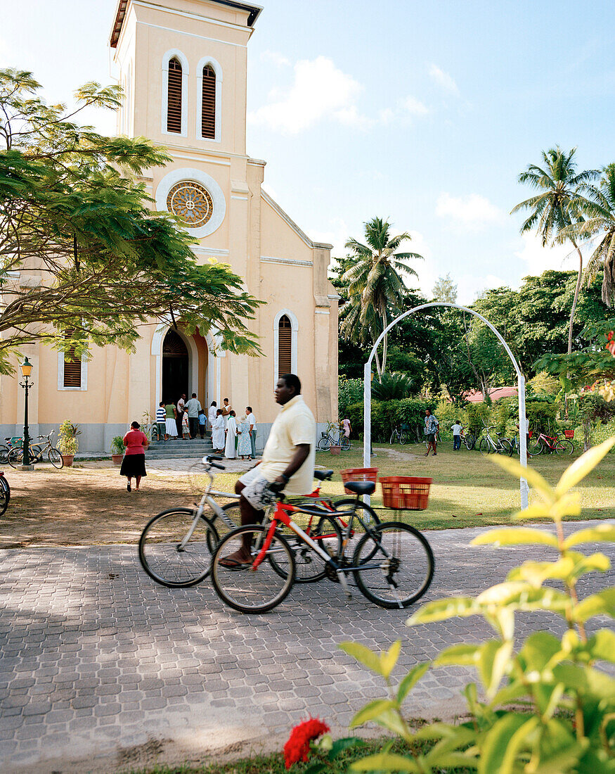 Man with rental bikes passing in front of the church at La Passe, La Digue, La Digue and Inner Islands, Republic of Seychelles, Indian Ocean
