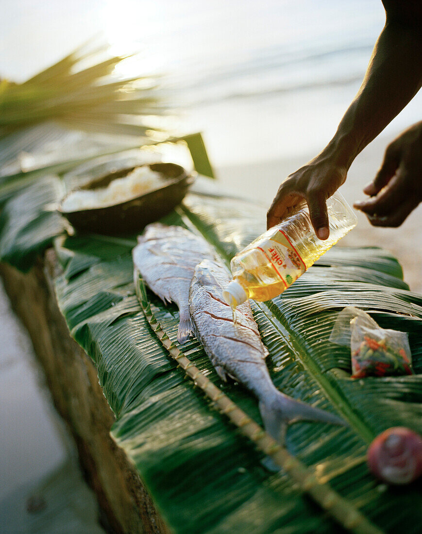 Mann bereitet Fisch auf kreolische Art zum Grillen vor, Strand von La Passe, im Hintergrund Nachbarinsel Praslin,  La Digue, La Digue and Inner Islands, Republik Seychellen, Indischer Ozean