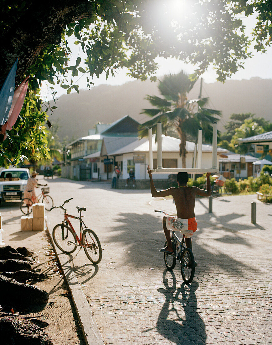 Biker carrying plastic table on his head on main road in the morning, La Passe, La Digue and Inner Islands, Republic of Seychelles, Indian Ocean