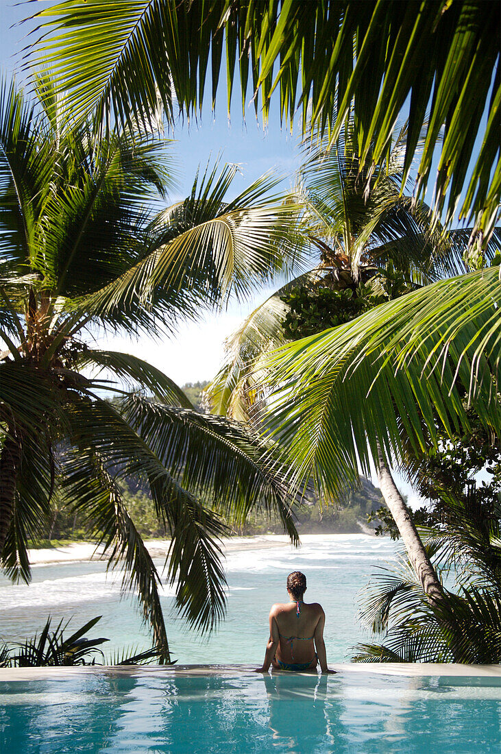 Woman sitting on edge of infinity pool overlooking coastline, North Island, Seychelles