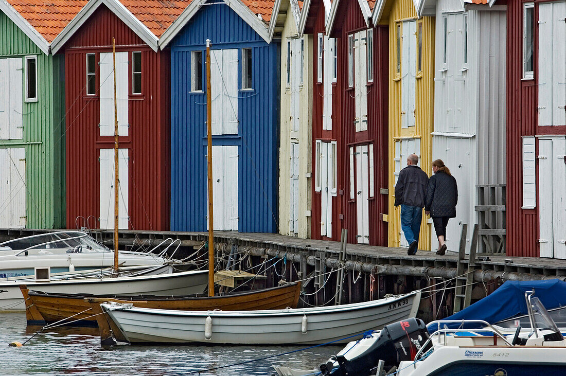 A couple strolling past the colourful fishing huts along the quayside at Smogen, Sotenas Municipality, Sweden.